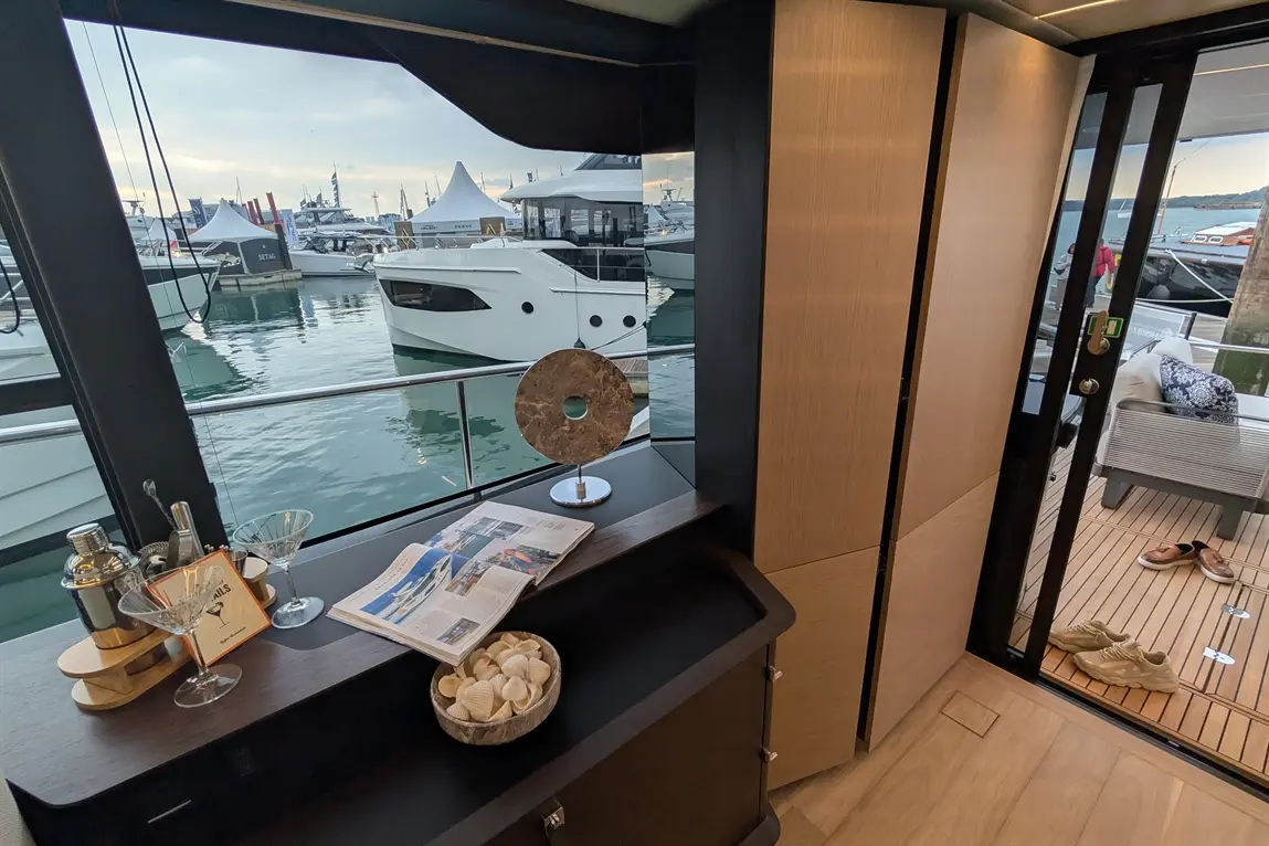 Elegant interior of a yacht showing a view of moored boats, a cocktail setup, seashells, and a newspaper on a counter.
